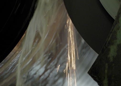 Schleifvorgang einer Metalloberfläche mit einer rotierenden Schleifscheibe, wobei Funken und Schleifflüssigkeit spritzen.